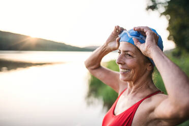 Cheerful senior woman in swimsuit standing by lake outdoors before swimming. - HPIF18956