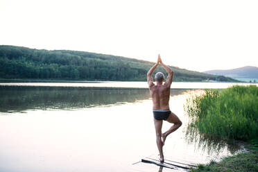 Rear view of senior man in swimsuit standing by lake outdoors doing yoga. Copy space. - HPIF18971