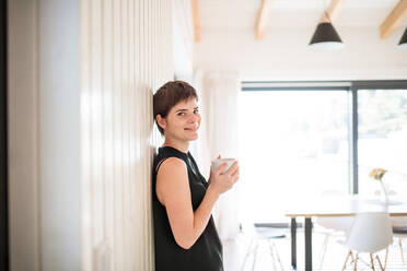 A young woman with coffee standing indoors at home, leaning on wall. Copy space. - HPIF18985