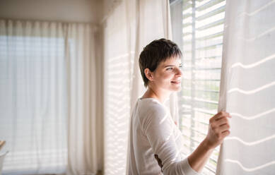 Portrait of young happy woman standing by window indoors at home, looking out through window. - HPIF18988