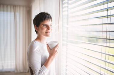 Porträt einer jungen glücklichen Frau mit Kaffee, die zu Hause am Fenster steht. - HPIF18990