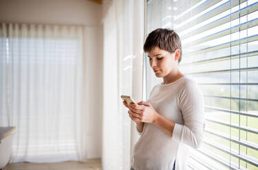 Porträt einer jungen Frau mit Smartphone, die zu Hause am Fenster steht und Textnachrichten schreibt. - HPIF18993