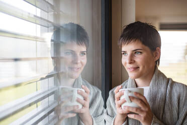 Porträt einer jungen glücklichen Frau mit Kaffee, die zu Hause am Fenster steht. - HPIF18996