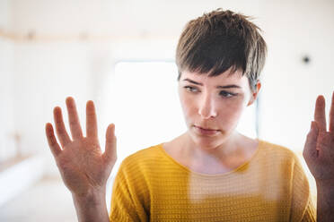 Front view of sad young woman standing indoors at home, hands on window. Shot through glass. - HPIF19001