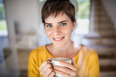 Front view of young woman with coffee standing indoors at home. Shot through glass. - HPIF19009