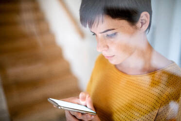 Eine junge Frau mit Smartphone steht in einem Haus, aufgenommen durch Glas. - HPIF19015