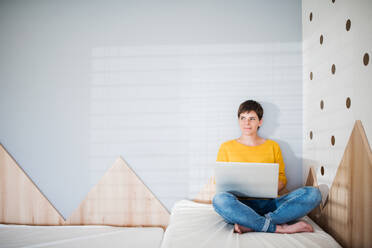 Vorderansicht einer jungen Frau mit Laptop, die auf dem Bett im Schlafzimmer zu Hause sitzt, Kopierraum. - HPIF19022