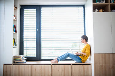 Seitenansicht einer jungen Frau mit Laptop, die auf der Fensterbank im Schlafzimmer zu Hause sitzt. - HPIF19034