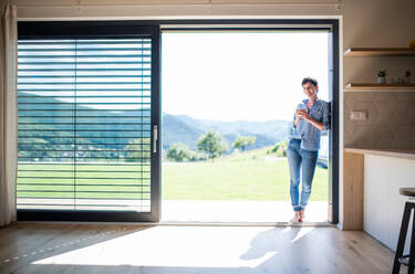 Front view of young woman with coffee standing by patio door at home. Copy space. - HPIF19035