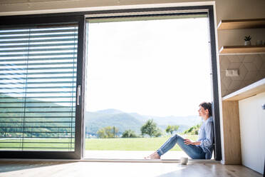 Seitenansicht einer jungen Frau mit Kaffee, die zu Hause an der Terrassentür sitzt, Raum kopieren. - HPIF19039