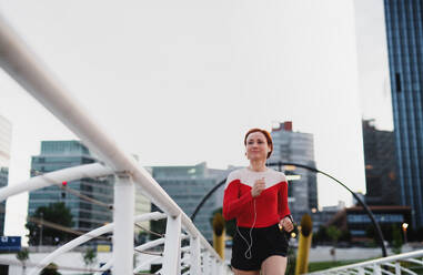 Front view of young woman runner with earphones jogging outdoors in city. - HPIF19136