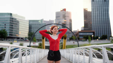 Front view of young woman runner with earphones in city, resting on the bridge. - HPIF19139