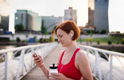 Eine junge Frau ruht sich nach einer Übung auf einer Brücke im Freien in der Stadt aus und benutzt ihr Smartphone. - HPIF19163