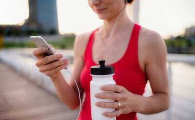 Mittelteil einer jungen Läuferin mit Kopfhörern in der Stadt, die beim Ausruhen ihr Smartphone benutzt. - HPIF19164