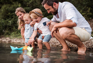Eine junge Familie mit zwei Kleinkindern, die im Sommer draußen am Fluss mit Papierbooten spielen. - HPIF19225