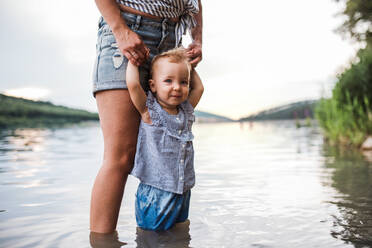 A midsection of mother with a toddler daughter outdoors by the river in summer, standing in water. - HPIF19230