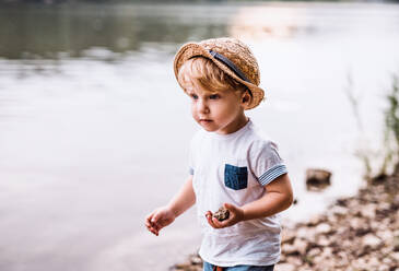 A small toddler boy standing barefoot outdoors by a river in summer, playing with rocks. - HPIF19235