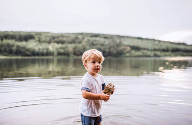 Ein nasses, kleines Kleinkind steht im Sommer barfuß in einem Fluss und spielt mit Steinen. - HPIF19237