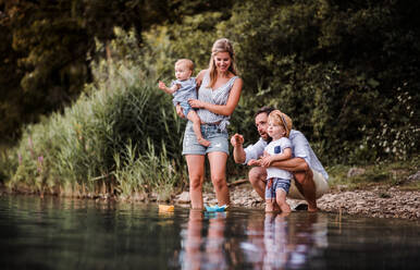 A young family with two toddler children outdoors by the river in summer, playing with paper boats. - HPIF19255