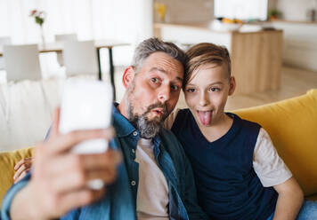 Mature father with small son sitting on sofa indoors, grimacing when taking selfie. - HPIF19460