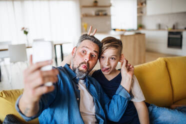 Mature father with small son sitting on sofa indoors, grimacing when taking selfie. - HPIF19461