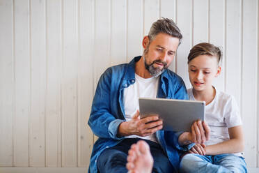 Mature father with small son sitting indoors against white wall, using tablet. Copy space. - HPIF19468
