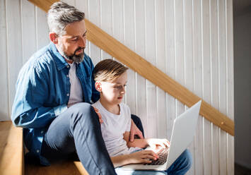 Ein reifer Vater mit kleinem Sohn sitzt auf der Treppe in einem Haus und benutzt einen Laptop. - HPIF19471