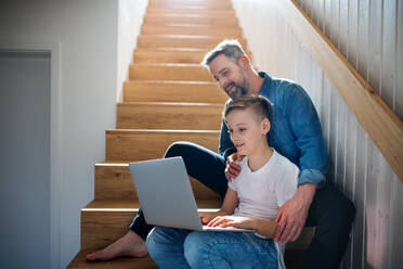 A mature father with small son sitting on the stairs indoors, using laptop. - HPIF19472