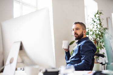 Geschäftsmann im Rollstuhl im Büro, mit einer Tasse Kaffee in der Hand. - HPIF19481