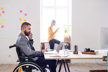 Zwei Geschäftsleute, die im Büro zusammenarbeiten. Ein Mann im Rollstuhl mit einem Smartphone, der einen Anruf tätigt. - HPIF19494