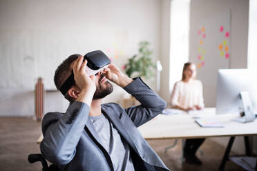 Zwei Geschäftsleute mit Rollstuhl im Büro arbeiten zusammen. Ein Mann trägt eine VR-Brille. - HPIF19508