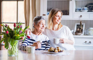 Porträt einer älteren Großmutter mit einer erwachsenen Enkelin zu Hause. Die Frauen sitzen am Tisch, essen Kuchen und trinken Kaffee. - HPIF19607