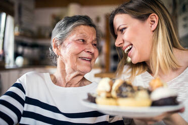 Porträt einer älteren Großmutter mit einer erwachsenen Enkelin zu Hause. Die Frauen sitzen am Tisch und halten einen Teller voller Kuchen. - HPIF19609