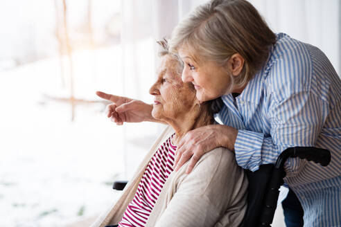 Eine ältere Mutter mit ihrer Tochter zu Hause, die aus dem Fenster schaut. Eine Pflegekraft, die einer behinderten älteren Frau im Rollstuhl hilft. - HPIF19743