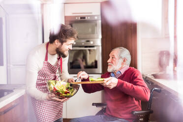 Junger Sohn mit seinem älteren Vater im Rollstuhl beim Kochen in der Küche. Zwei Generationen in einem Haus. Aufnahme durch Glas. - HPIF19853