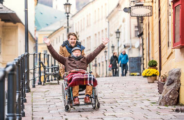 Ein Vater im Rollstuhl und sein kleiner Sohn gehen spazieren und haben Spaß. Ein Pfleger hilft einem behinderten älteren Mann. - HPIF19895
