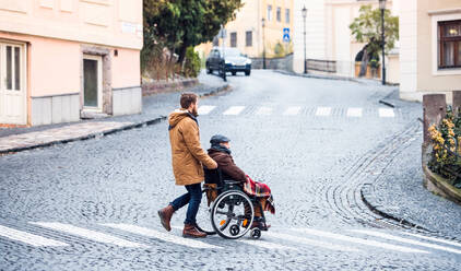 Father in wheelchair and young son on a walk, crossing the road. A carer assisting disabled senior man. - HPIF19897