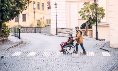 Father in wheelchair and young son on a walk., crossing the road. A carer assisting disabled senior man. - HPIF19898