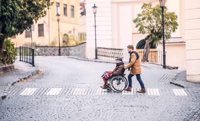 Vater im Rollstuhl und kleiner Sohn beim Spazierengehen, der die Straße überquert. Ein Pfleger hilft einem behinderten älteren Mann. - HPIF19898