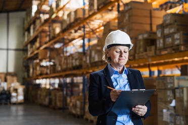 Senior woman warehouse manager or supervisor with a white helmet and clipboard, controlling stock. - HPIF19933