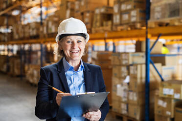 Senior woman warehouse manager or supervisor with a white helmet and clipboard, controlling stock. - HPIF19934