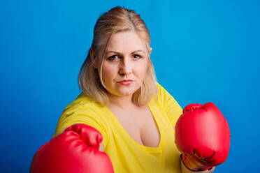 Porträt einer übergewichtigen Frau mit roten Boxhandschuhen im Studio, Studioaufnahme vor blauem Hintergrund. - HPIF20115