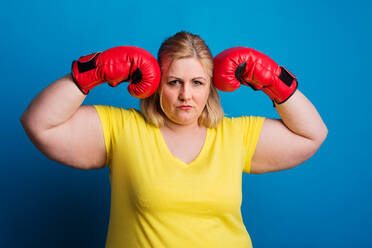 Porträt einer übergewichtigen Frau mit roten Boxhandschuhen im Studio, Studioaufnahme vor blauem Hintergrund. - HPIF20116