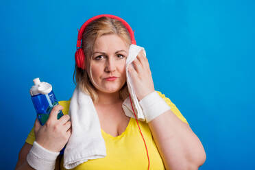 Porträt einer übergewichtigen Frau mit Kopfhörern und Wasserflasche im Studio auf blauem Hintergrund, die sich das Gesicht mit einem Handtuch abwischt. Eine Studioaufnahme auf blauem Hintergrund. - HPIF20117