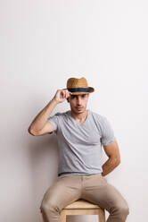 Ein selbstbewusster hispanischer junger Mann mit Hut sitzt auf einem Hocker in einem Studio und hält eine Krempe. - HPIF20129