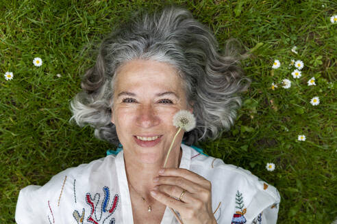 Smiling woman holding dandelion blowball lying on green grass - FLLF00892