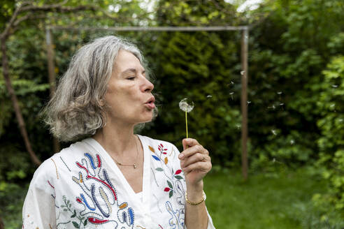 Ältere Frau bläst Pusteblumen im Garten - FLLF00893