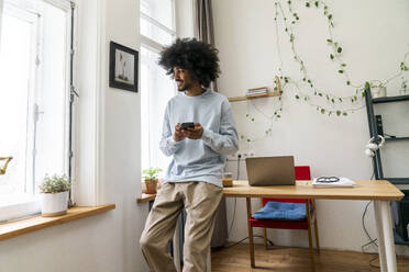 Smiling freelancer holding smart phone leaning on desk at home - VPIF08164