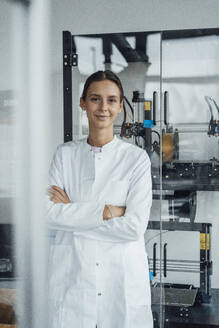 Smiling young scientist standing with arms crossed in front of glass cabinet - JOSEF19557