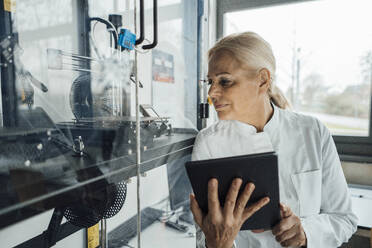 Senior scientist with tablet PC examining robotic arm in glass cabinet - JOSEF19582